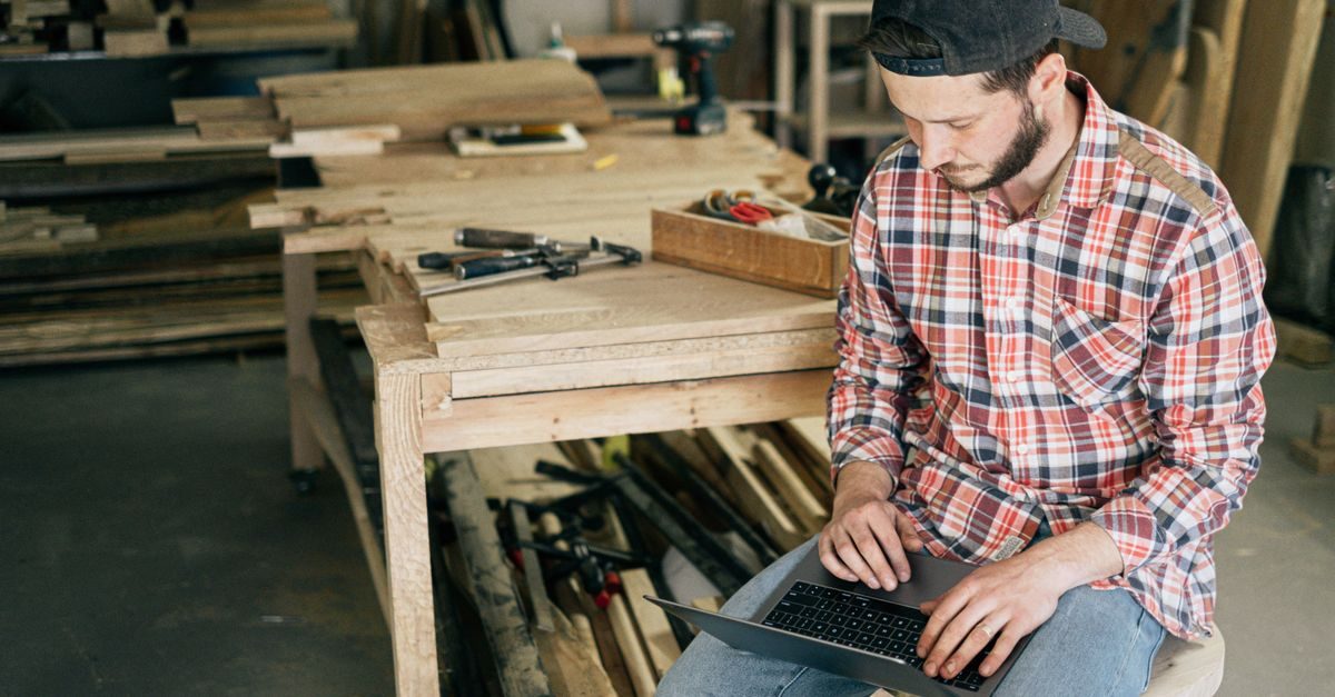 Falegname con camicia a quadri lavora concentrato al computer portatile in una bottega artigiana tra legno e attrezzi