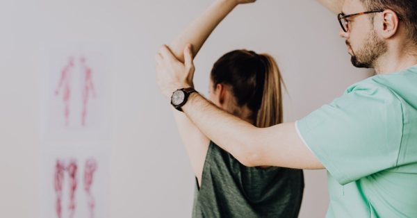 A male physiotherapist in a green scrub top examines a female patient's shoulder in a clinical setting