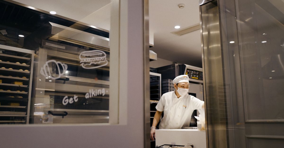 Bakker met mondkapje en koksmuts werkt in een professionele keuken achter een glazen wand met getekende iconen