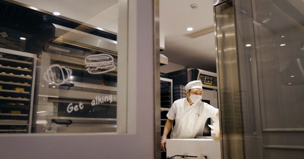 Bakker met mondkapje en koksmuts werkt in een professionele keuken achter een glazen wand met getekende iconen