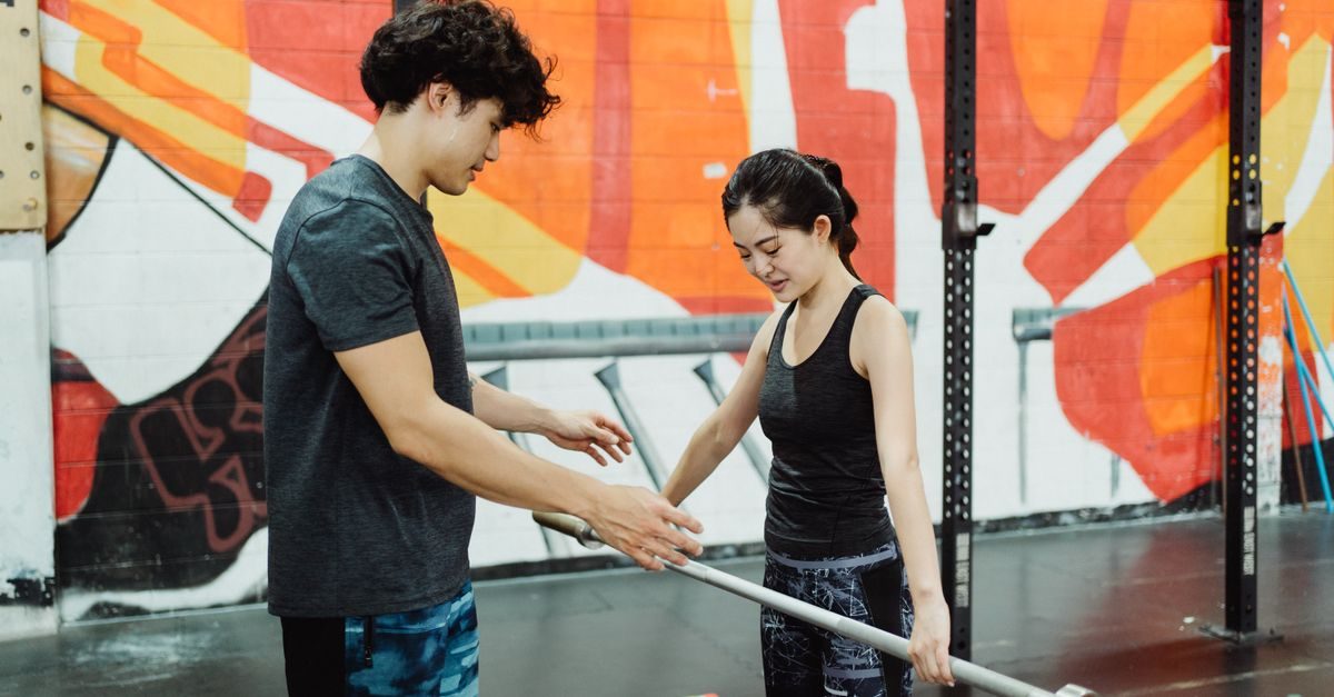 Un coach sportif conseille une femme tenant une barre de musculation dans une salle de sport au mur coloré