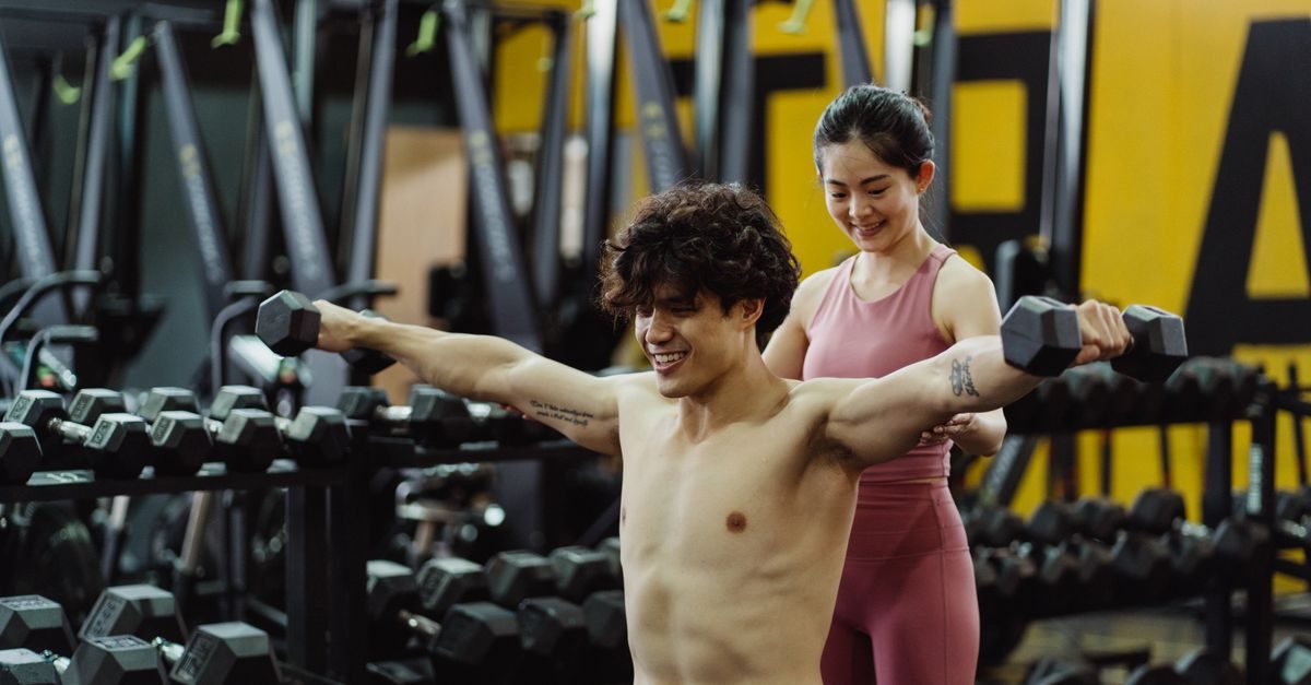 En leende man tränar axlar med hantlar på gymmet medan en kvinnlig instruktör ger stöd och coachning