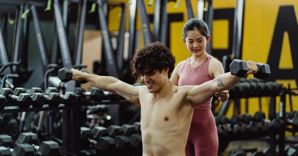 En shirtlös man med lockigt hår lyfter hantlar med armarna utsträckta åt sidorna medan en kvinna i rosa träningsställ hjälper honom i ett gym