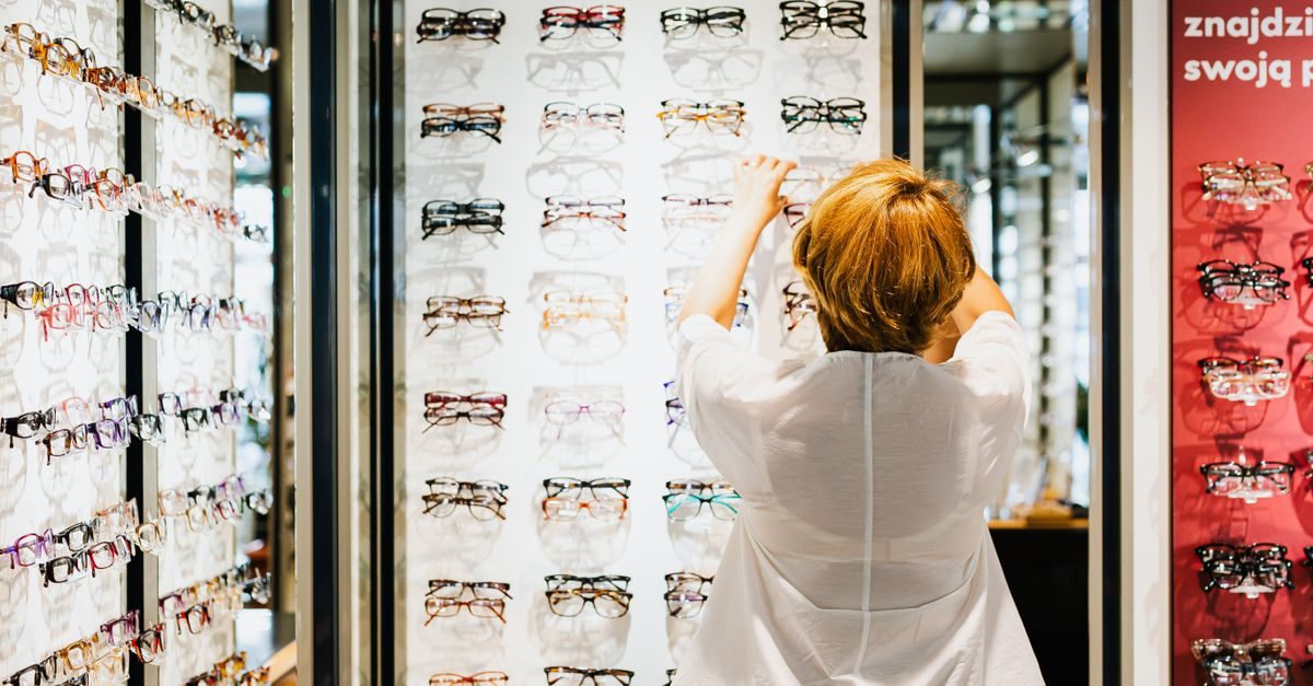 Kobieta w białej bluzce wybiera okulary z szerokiej oferty prezentowanej na podświetlanej ścianie w salonie optycznym