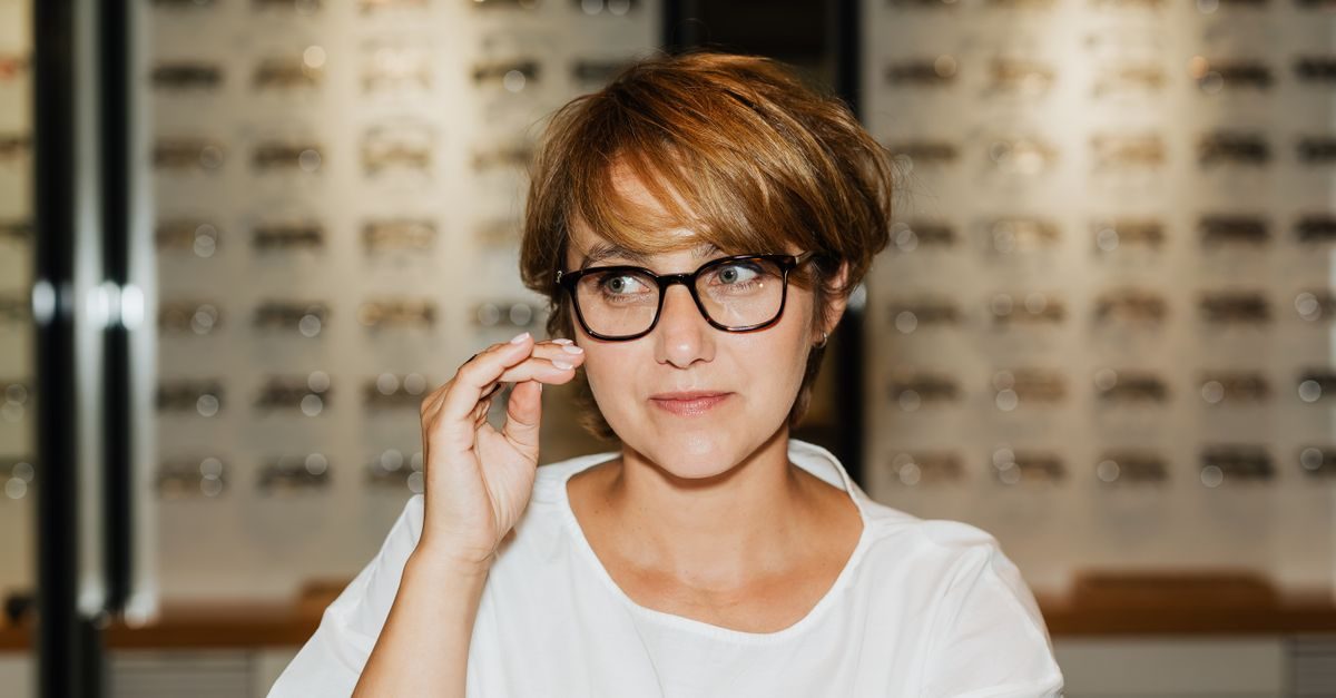 Kvinna med kort hår provar svarta glasögon i en optikbutik med väggar fyllda av glasögonbågar i bakgrunden
