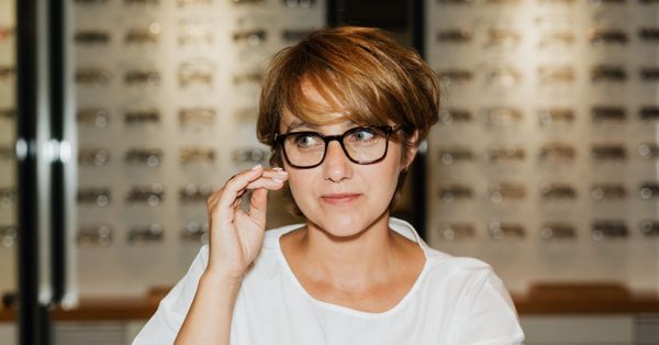 Kvinna med kort hår provar svarta glasögon i en optikbutik med väggar fyllda av glasögonbågar i bakgrunden