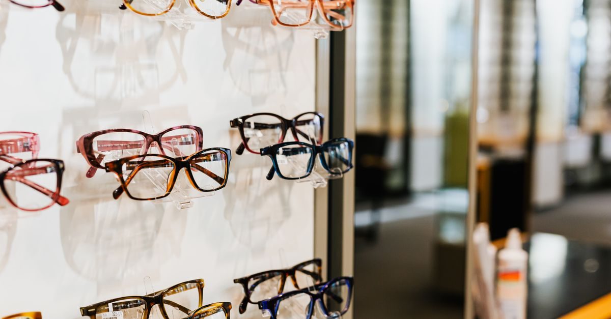 Mehrere Brillenmodelle in verschiedenen Farben und Formen hängen ordentlich präsentiert an einer weißen Wand im Optikergeschäft