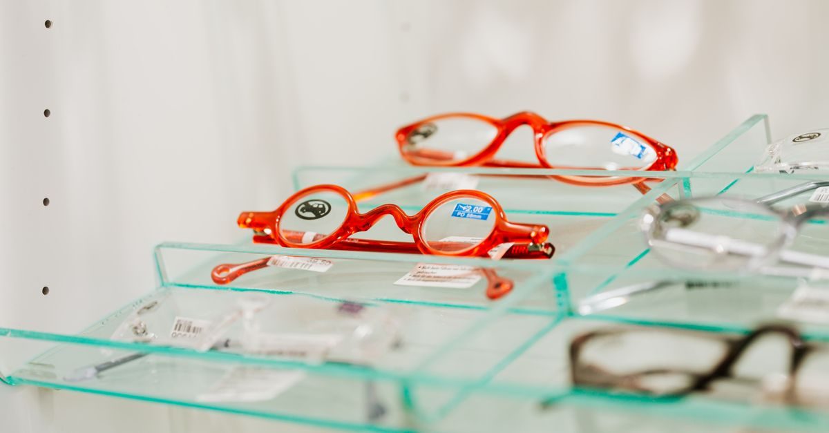 Bright orange eyeglasses sit on a glass display shelf in an optician's office with other frames visible nearby