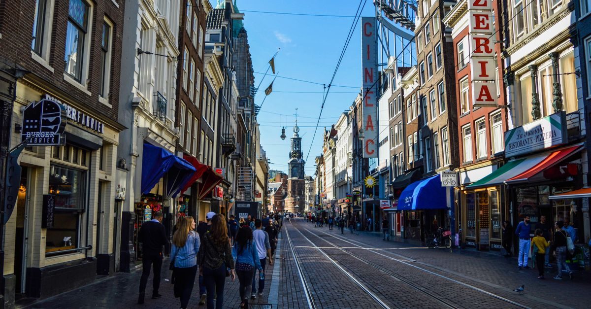 Mensen lopen door een drukke winkelstraat in Amsterdam met historische panden en uithangborden onder een heldere lucht