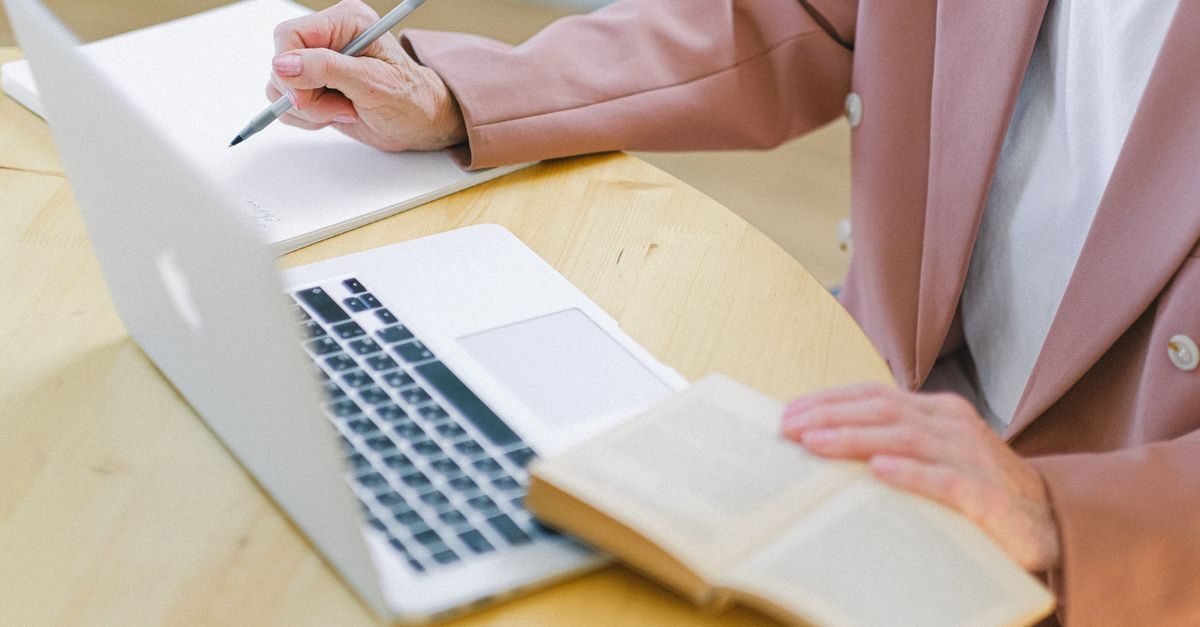 Iemand in een roze jasje schrijft met een pen in een notitieblok naast een laptop op een houten tafel