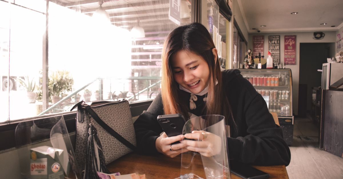 Eine lächelnde junge Frau sitzt in einem Café und schaut auf ihr Smartphone