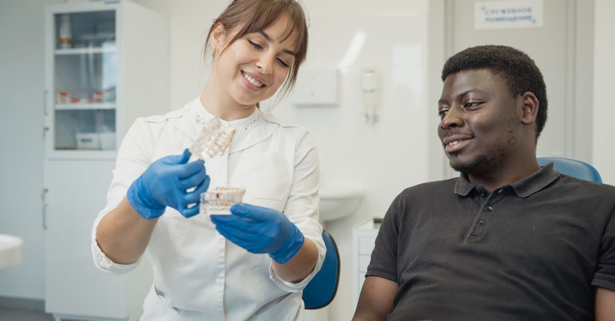 A smiling dentist in a white coat and blue gloves explains a dental model to a patient in a bright clinical office