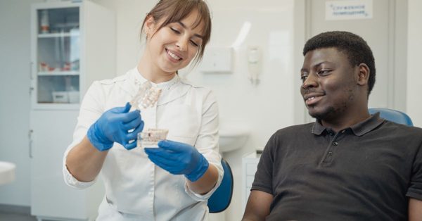 A smiling dentist in a white coat and blue gloves explains a dental model to a patient in a bright clinical office