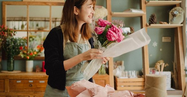 Eine lächelnde Floristin verpackt in ihrem gemütlichen Blumenladen einen Strauß pinker Rosen in helles Papier