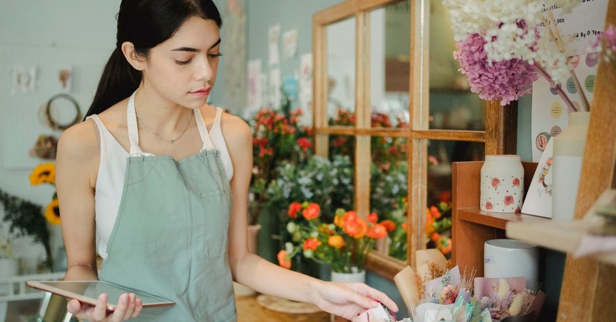 Jonge bloemist met schort houdt een tablet vast en schikt bloemen in een sfeervolle winkel