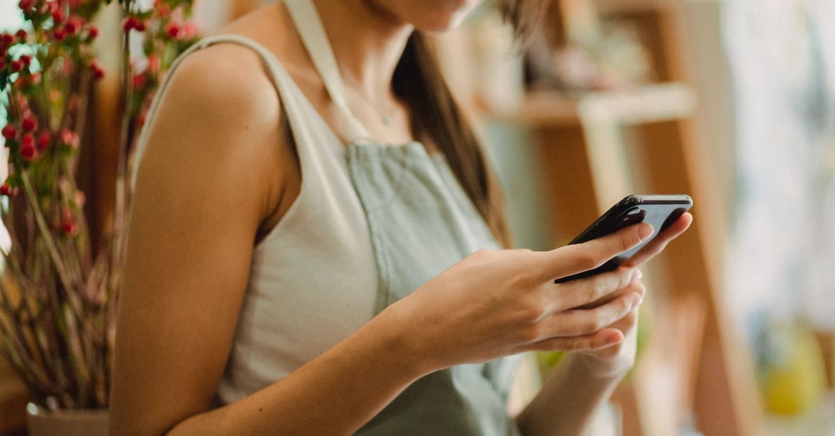 Eine Frau in einer hellen Schürze hält konzentriert ein Smartphone in den Händen, im Hintergrund steht eine Pflanze