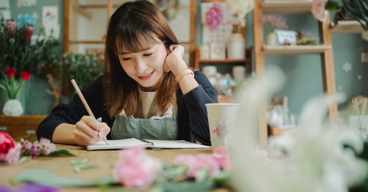 A smiling woman in an apron writes in a notebook at a wooden table surrounded by flowers in a cozy shop
