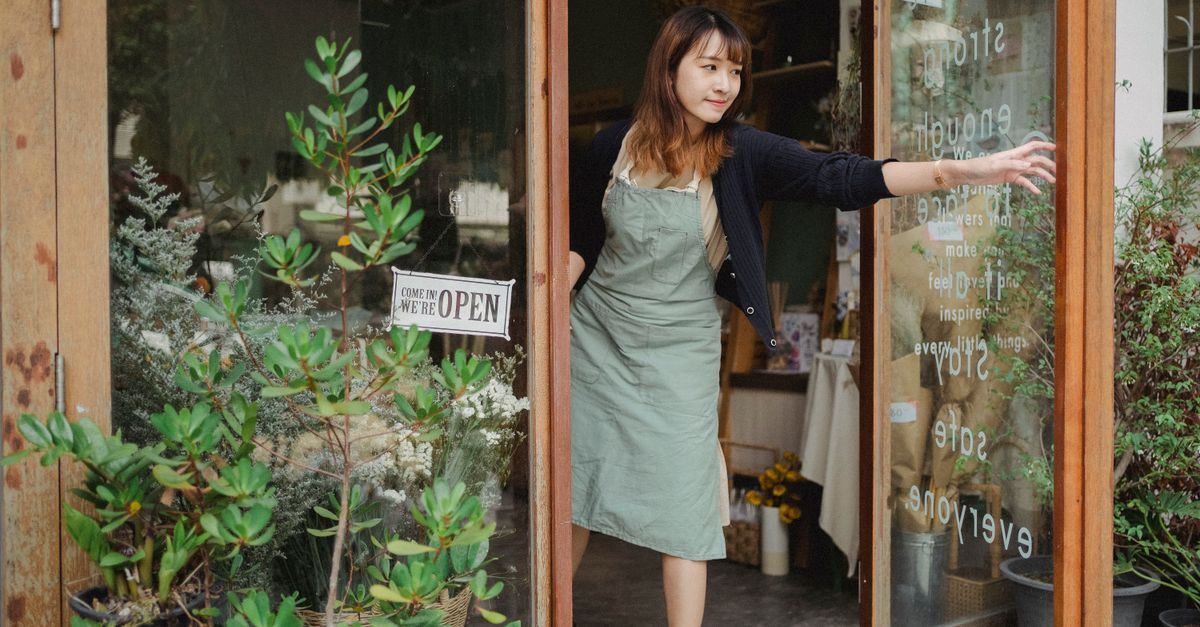 Une femme souriante en tablier ouvre la porte vitrée d'une boutique accueillante avec un panneau ouvert