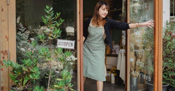 Une femme souriante en tablier ouvre la porte vitrée d'une boutique accueillante avec un panneau ouvert