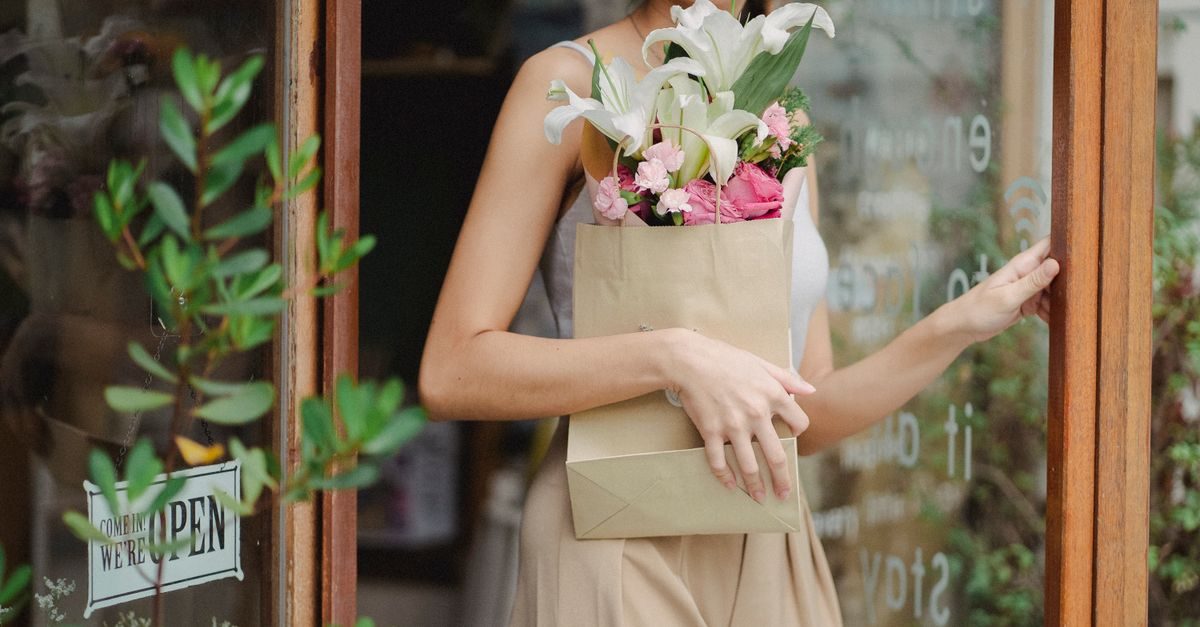 A woman in a light top exits a flower shop carrying a paper bag filled with white lilies and pink roses