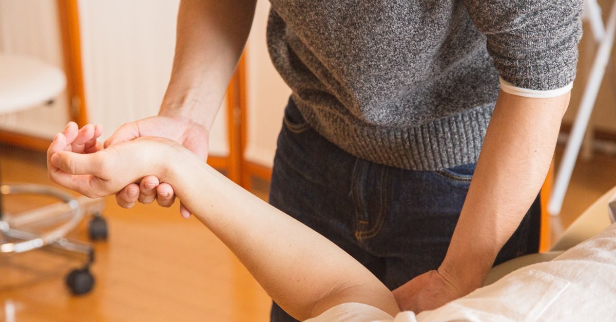 Terapeut i grå tröja behandlar en patients handled och underarm på britsen i ett ljust behandlingsrum
