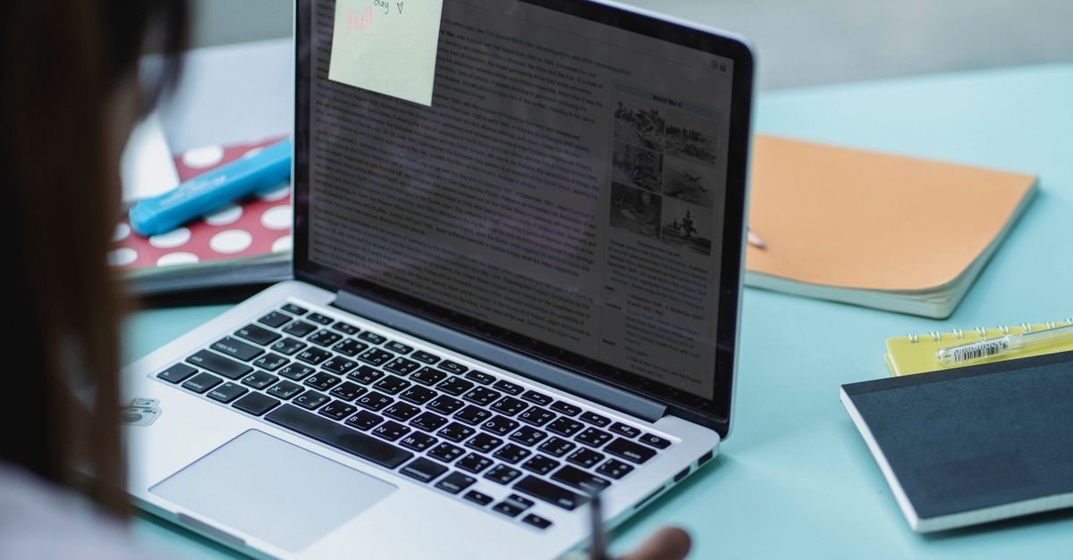 A person works on a laptop displaying a document with a sticky note on a desk cluttered with notebooks and stationery