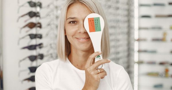 Mujer sonriente en una óptica cubriéndose un ojo con un oclusor de examen visual frente a estantes con gafas