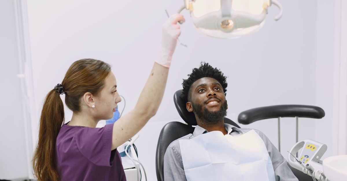 A female dentist in scrubs adjusts a bright overhead light for a smiling male patient sitting in a dental chair