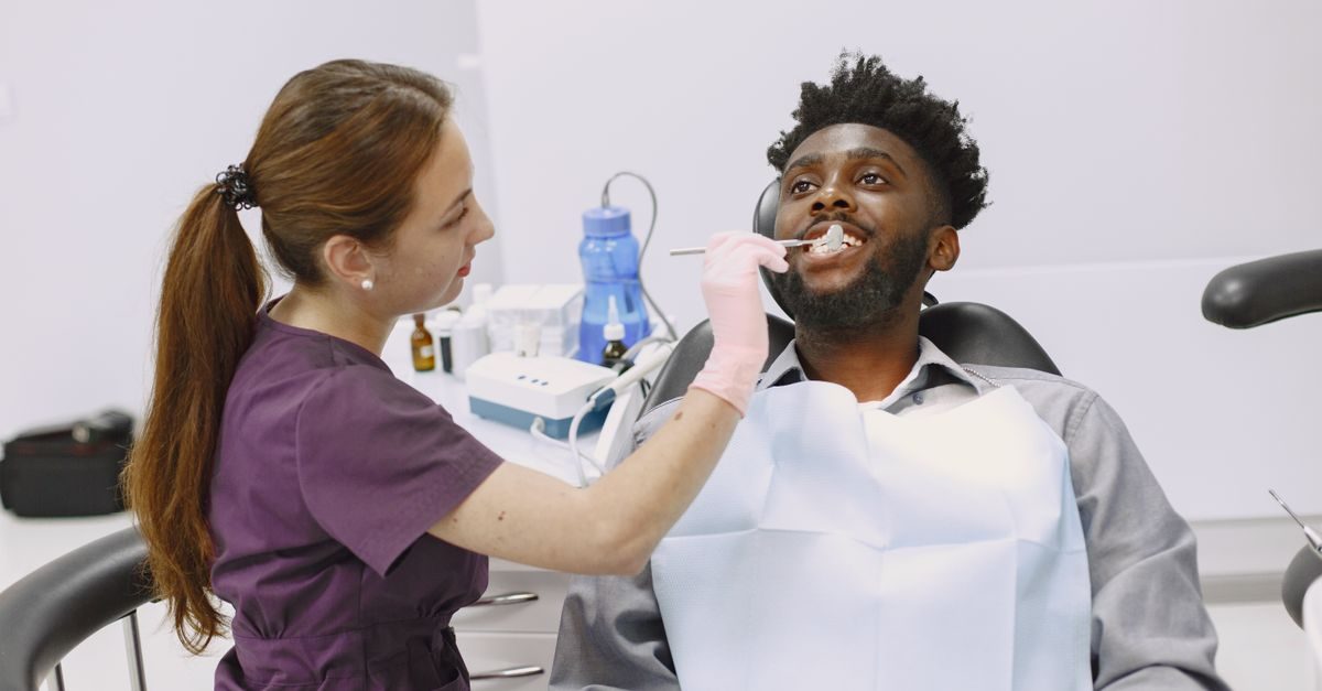 A female dentist in a purple uniform examines the teeth of a smiling male patient in a bright, modern dental clinic