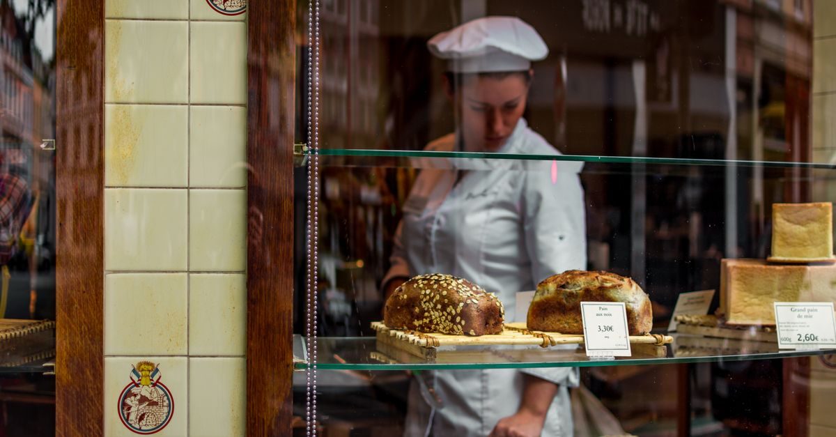 Eine Bäckerin in Arbeitskleidung steht konzentriert hinter einer gläsernen Verkaufstheke mit verschiedenen Brotsorten