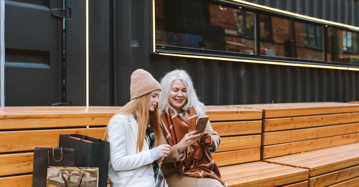 Twee vrouwen zitten met winkeltassen op een houten bank en kijken glimlachend samen naar een smartphone