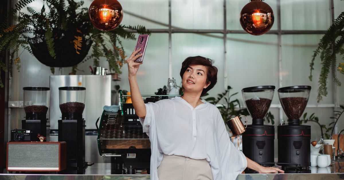 Une femme souriante prend un selfie avec son smartphone dans un café moderne entouré de plantes et de moulins à café