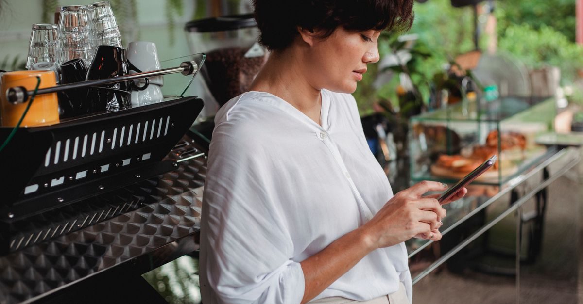 Vrouw in wit overhemd kijkt geconcentreerd op haar smartphone bij een espressomachine in een koffiebar