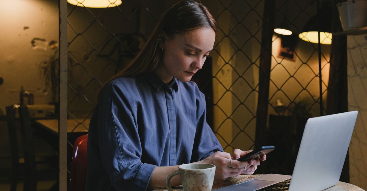 A focused woman in a blue shirt sits at a table in a dimly lit cafe, using her smartphone next to an open laptop