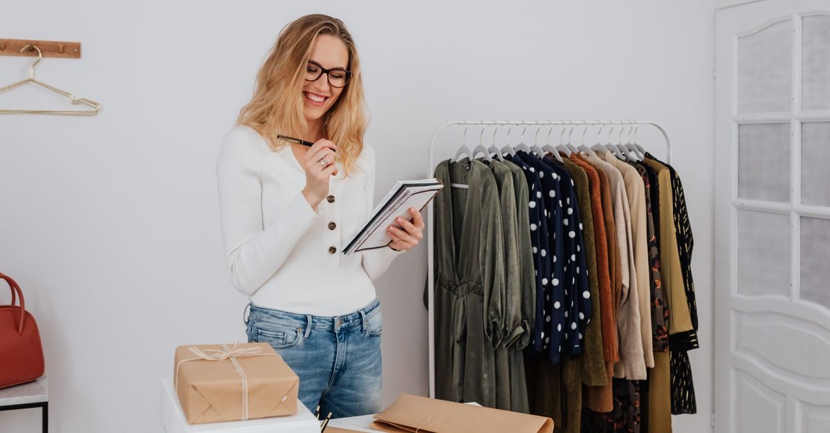 A smiling woman in a white top and glasses writes in a notebook near a clothing rack and wrapped packages in a studio