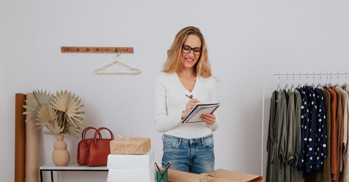 Une femme souriante à lunettes prend des notes dans un carnet au milieu d'un atelier de vêtements organisé