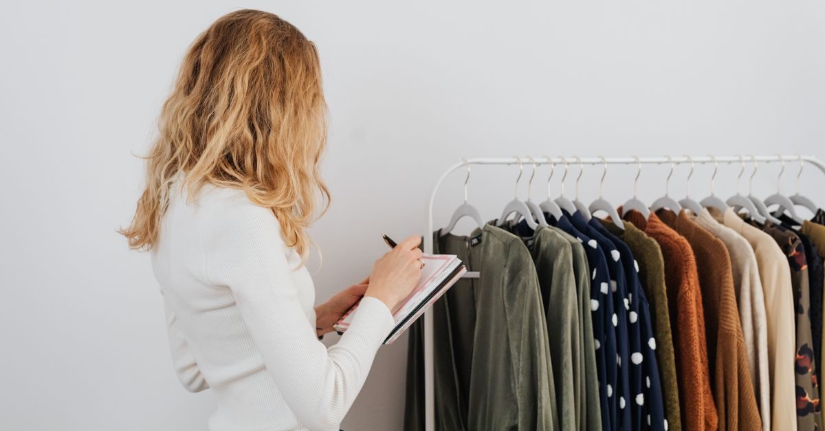 A woman with blonde hair stands in front of a clothing rack, focused on taking notes in a small notebook