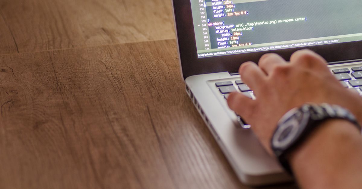 A person wearing a watch types code on a laptop screen while sitting at a wooden desk