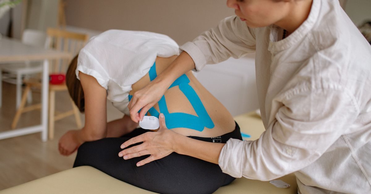 Eine Physiotherapeutin bringt blaue Kinesio-Tapes auf dem Rücken einer sitzenden Patientin an