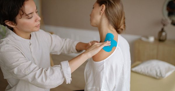 A physiotherapist applies blue kinesiology tape to a patient's shoulder in a calm clinic setting