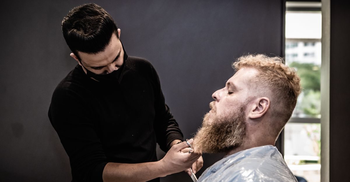 Fryzjer w czarnej koszulce strzyże nożyczkami brodę rudowłosego mężczyzny w profesjonalnym salonie fryzjerskim