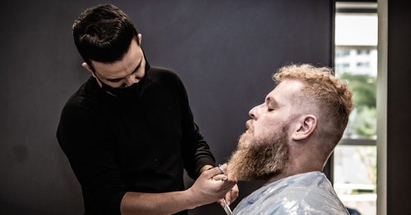 Fryzjer w czarnej koszulce strzyże nożyczkami brodę rudowłosego mężczyzny w profesjonalnym salonie fryzjerskim
