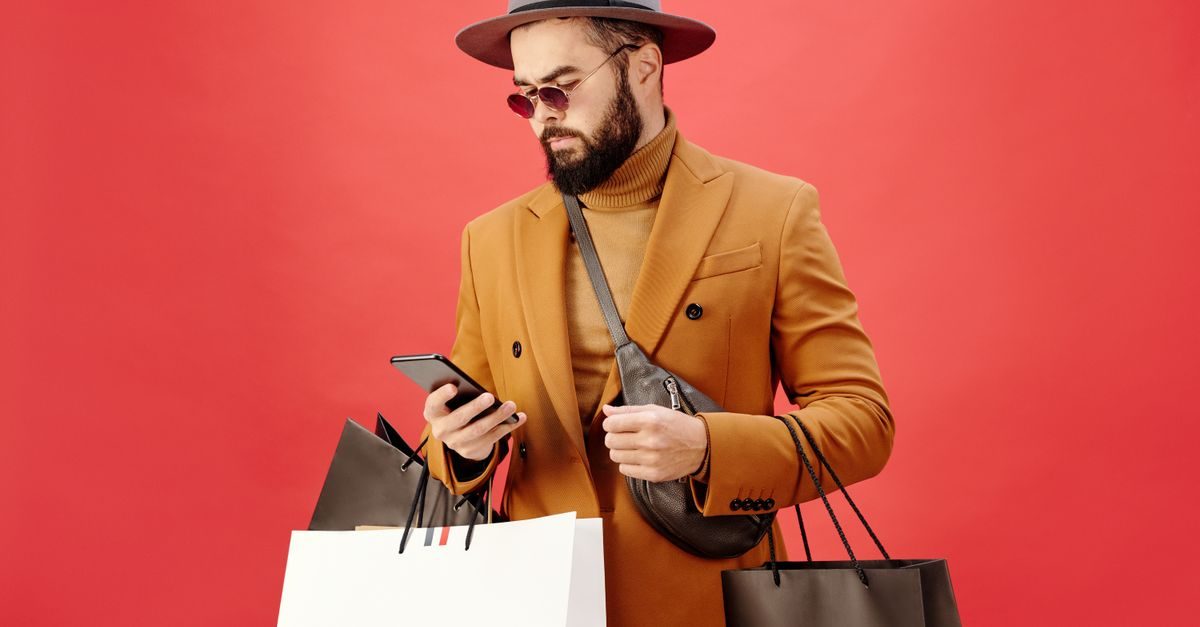 Stylish man in a tan coat, hat, and sunglasses looks at his smartphone while holding several shopping bags against red