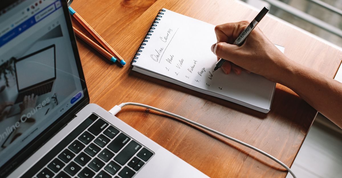 A person writes a list of online courses in a notebook next to a laptop on a wooden desk