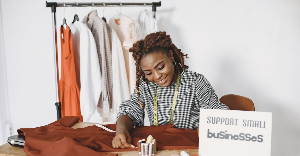 Glimlachende modeontwerpster werkt aan kledingstuk in haar atelier met een bordje met de tekst support small businesses