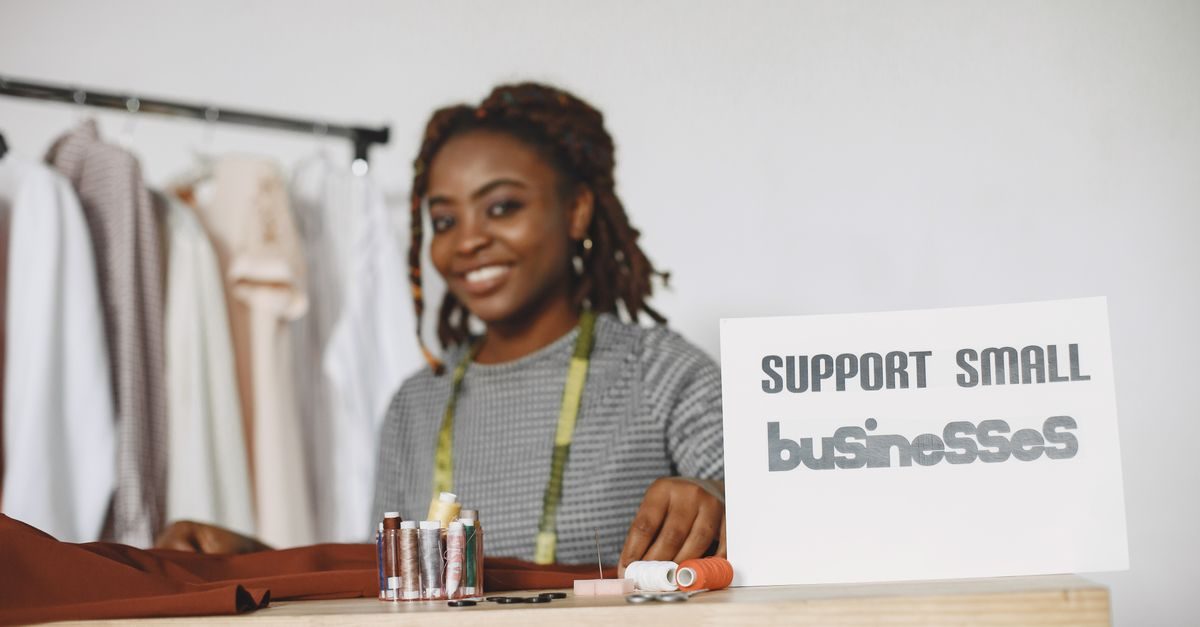 A smiling woman with dreadlocks works in her studio behind a sign that reads Support Small Businesses