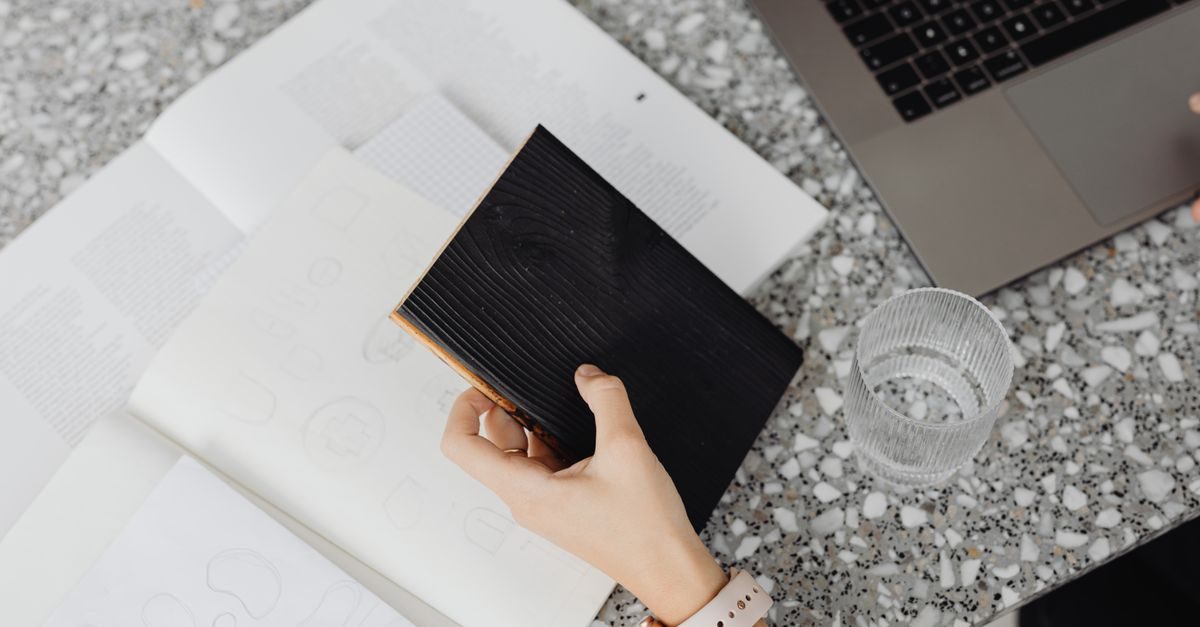 A person holds a black notebook over an open sketchbook next to a laptop and a glass of water on a speckled table