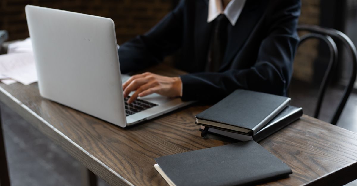 Persoon in zakelijk pak werkt op een laptop aan een houten bureau met notitieblokken in een kantooromgeving