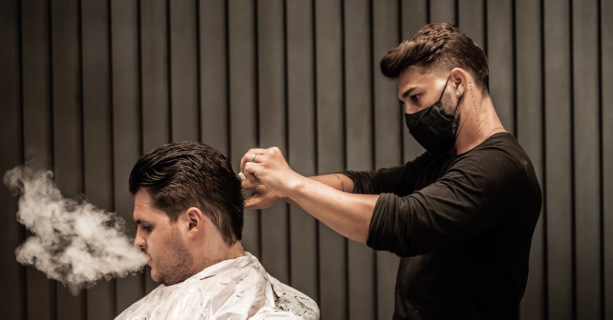 Barber w czarnej maseczce stylizuje włosy klienta, obok którego unosi się gęsta chmura pary na tle ciemnych listew ściennych