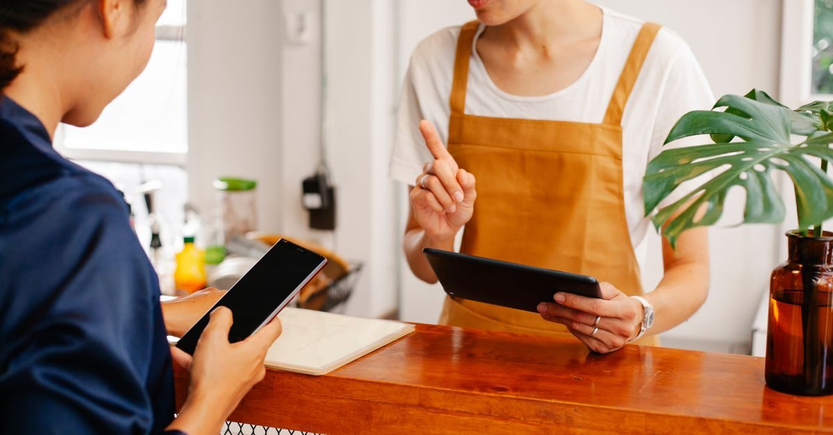Pracownik w fartuchu rozmawia z klientem przy ladzie, wskazując palcem na tablet w jasnym, nowoczesnym wnętrzu lokalu